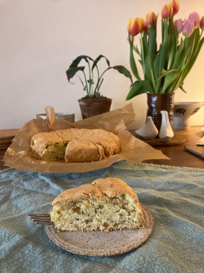 slice of seed cake on plate in foreground, cake in background with flowers