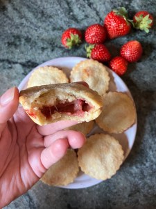 strawberry tart held by hand above plate with tarts, strawberries on counter